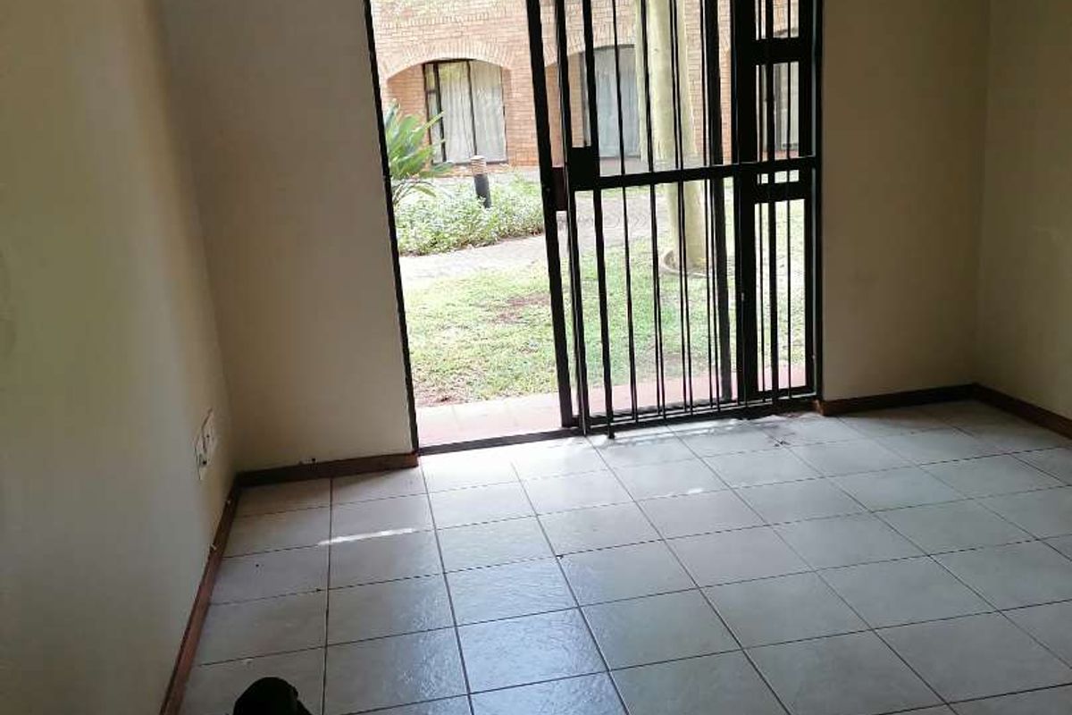 An empty living room featuring tiled flooring and a sliding glass door with a security gate, providing direct access to the outdoor garden area.