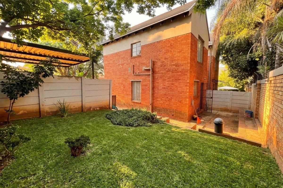 Rear view of a double-story face brick townhouse featuring a private garden, lawn, paved area, and a covered carport structure.