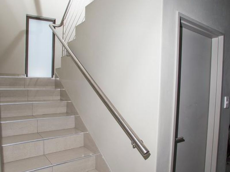 A bright, modern staircase featuring tiled steps, a sleek stainless steel handrail, and a tall vertical window providing natural light.