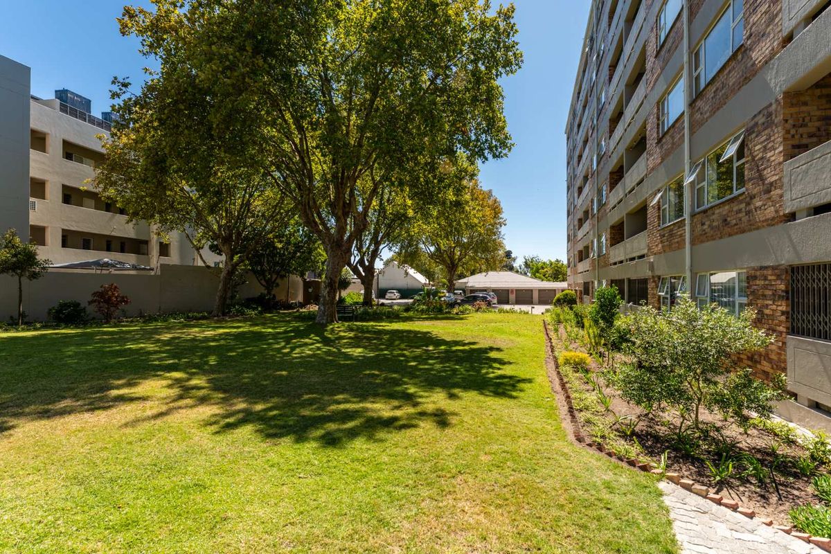 A well-maintained communal garden area featuring mature trees and landscaped borders adjacent to the apartment building exterior.