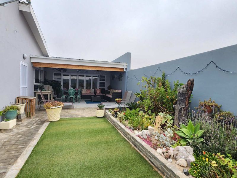 A well-maintained outdoor area featuring a covered patio with a built-in braai, brick paving, artificial lawn, and a landscaped raised garden bed.
