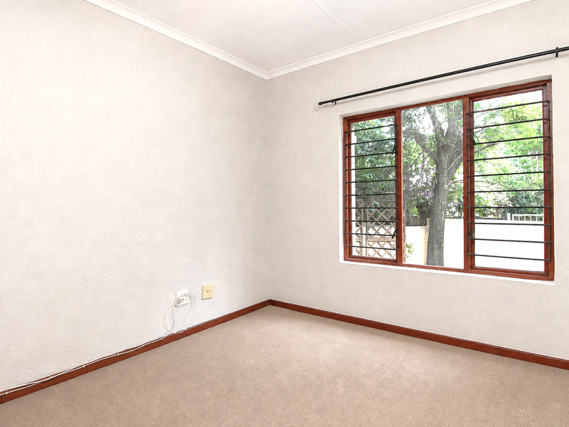 A clean, empty bedroom featuring neutral walls, carpeted flooring, and a large window with security bars, offering a blank canvas for personalization.