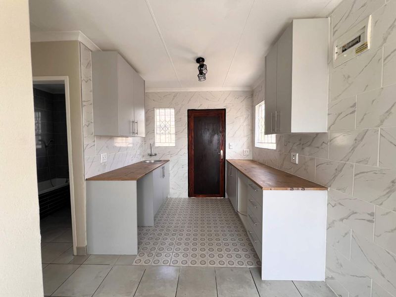 A clean, modern kitchen featuring light grey cabinetry, wooden countertops, marble-effect wall tiling, and tiled flooring, presented in an empty, move-in ready state.