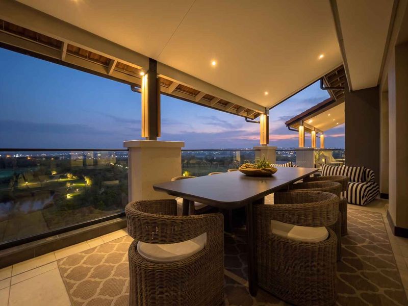 A spacious covered balcony featuring a glass balustrade, recessed lighting, and panoramic views, providing an ideal setting for outdoor dining and relaxation.