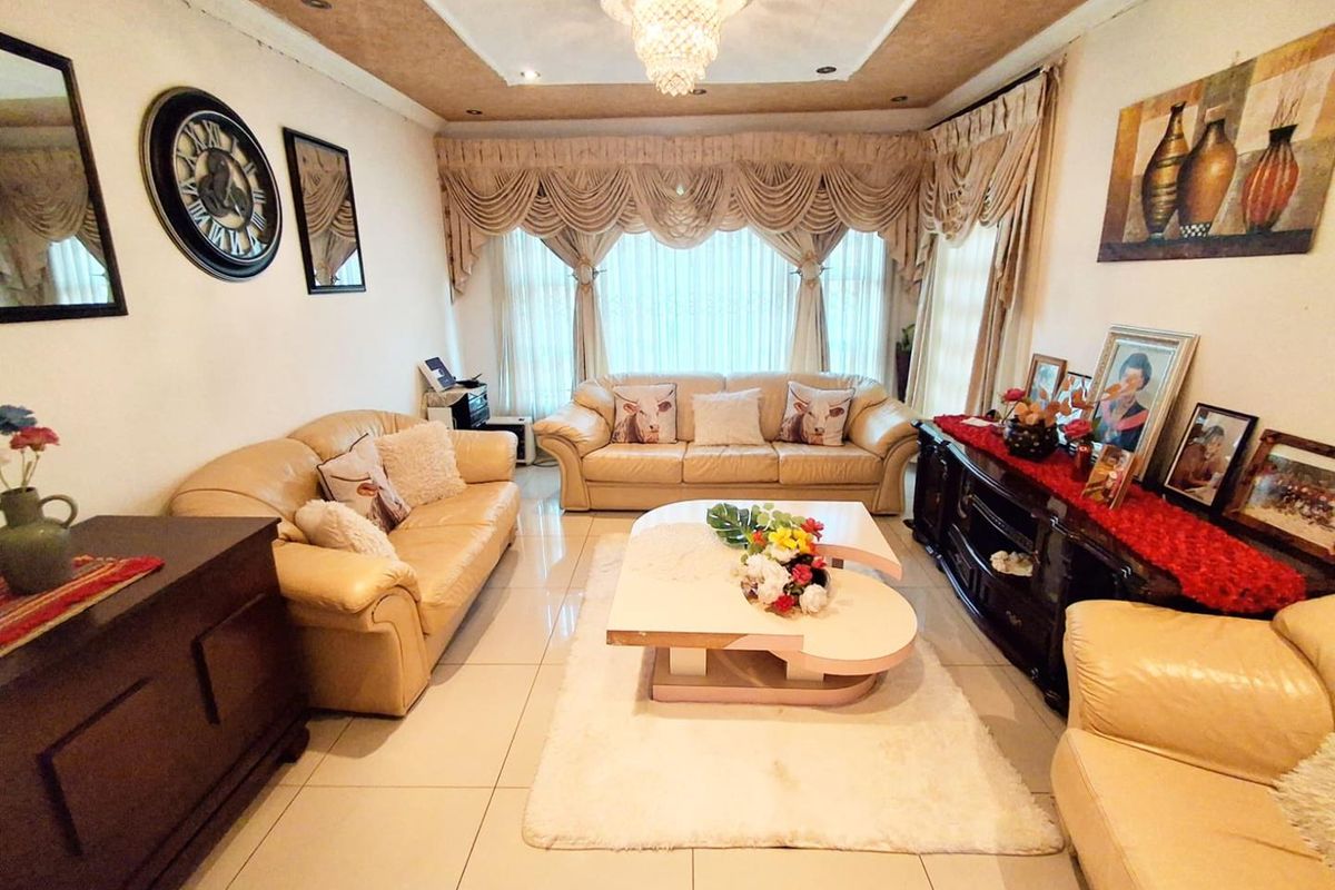 A spacious living room featuring tiled flooring, a decorative recessed ceiling with a central chandelier, and large windows with draped curtains.