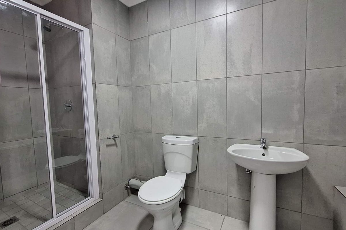 A modern bathroom featuring a glass-enclosed shower, pedestal basin, and toilet, finished with neutral grey wall and floor tiles.