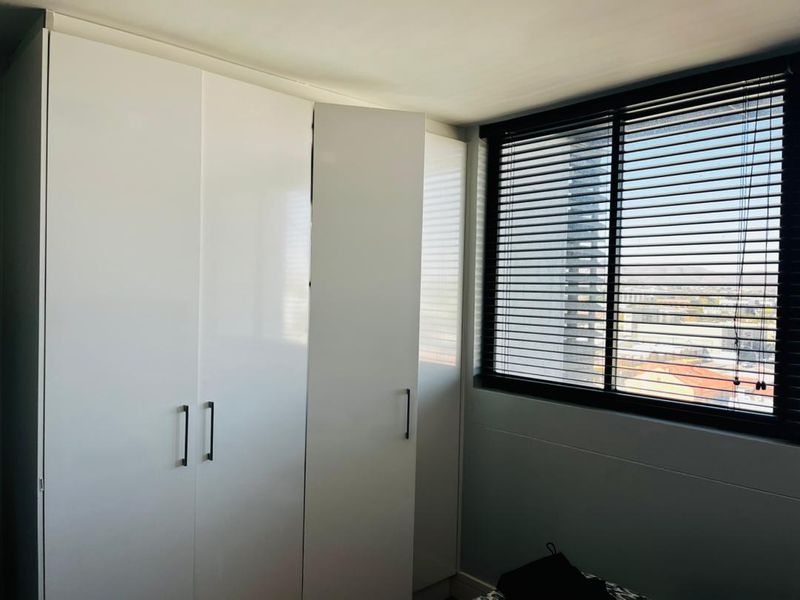 A bright bedroom featuring modern white built-in cupboards and dark venetian blinds, offering a clean and functional space with an urban view.