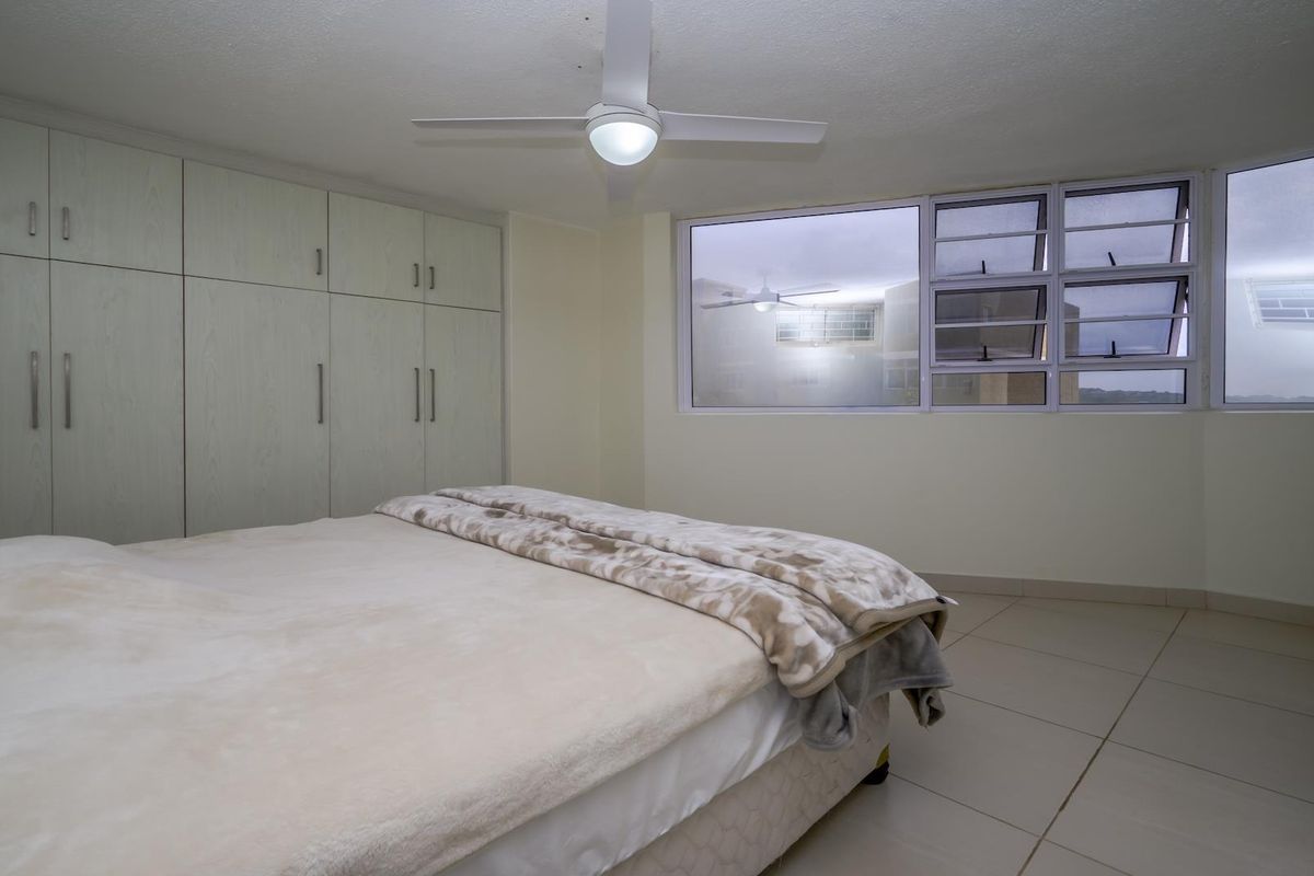 A bedroom featuring built-in cupboards, tiled flooring, and a ceiling fan, with large windows providing natural light.