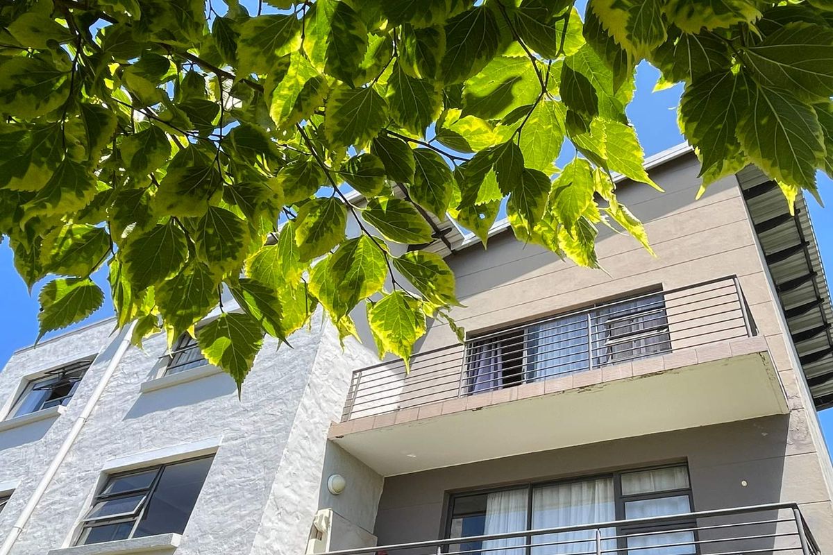 Exterior view of a multi-story apartment building featuring balconies with metal railings and modern facade finishes, partially framed by lush green foliage.