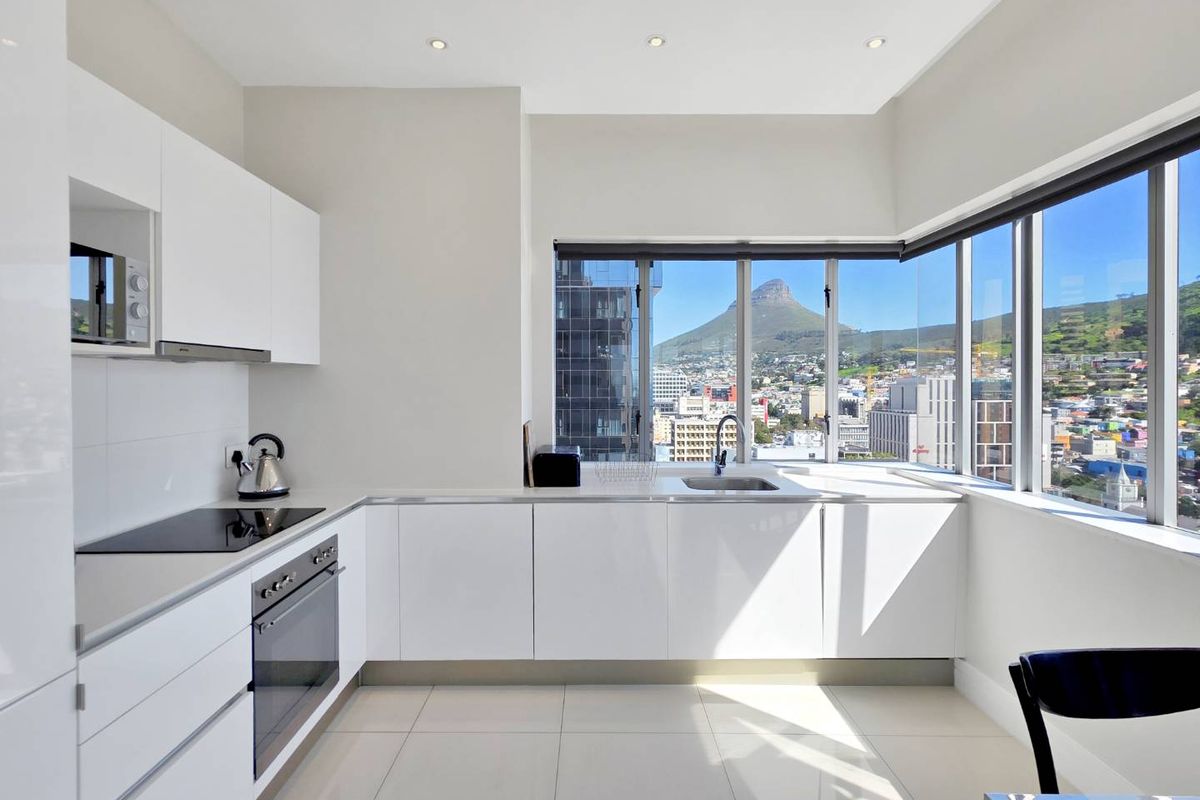 Modern kitchen featuring sleek white cabinetry, integrated appliances, tiled floors, and expansive corner windows offering stunning city and mountain views.