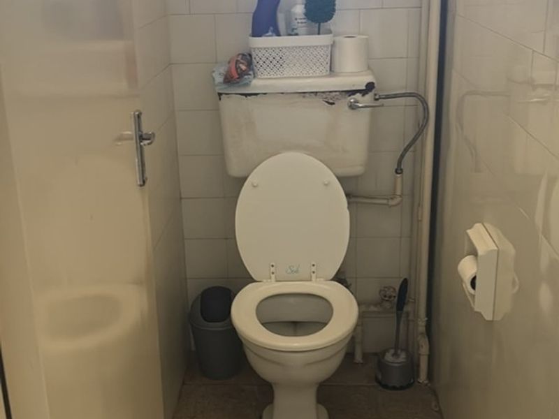 A compact bathroom featuring a standard toilet with a wall-mounted cistern, tiled walls, and tiled flooring, requiring cosmetic updates and maintenance.