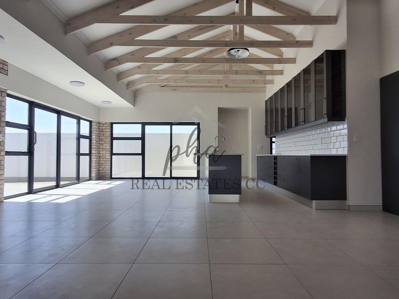 Spacious open-plan living and kitchen area featuring exposed timber trusses, modern dark cabinetry, tiled floors, and large aluminium sliding doors leading outdoors.