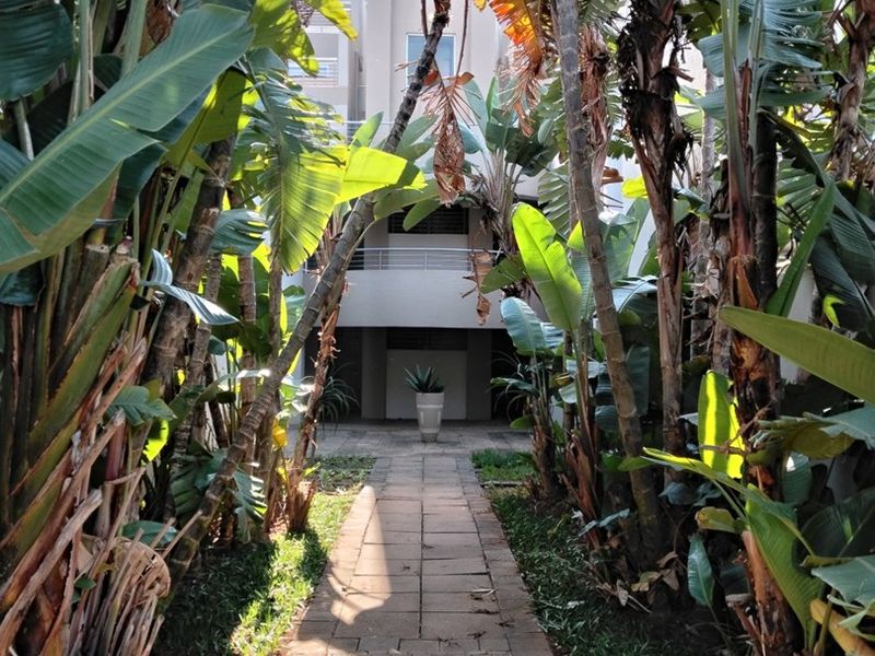 A paved garden walkway leading towards the rear facade of an apartment building, framed by mature tropical landscaping.