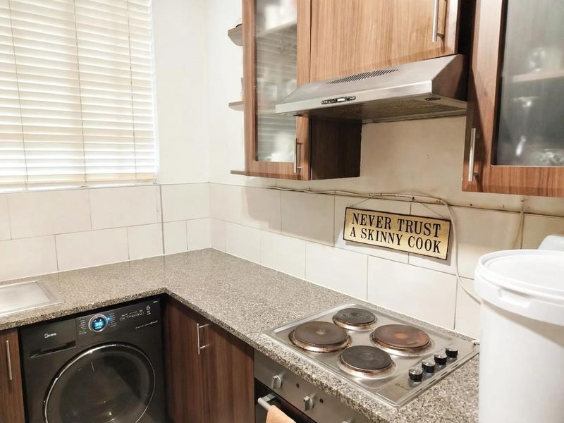 Compact kitchen featuring wood-finish cabinetry, granite countertops, an integrated electric hob, oven, extractor fan, and a stainless steel sink with tiled backsplash.