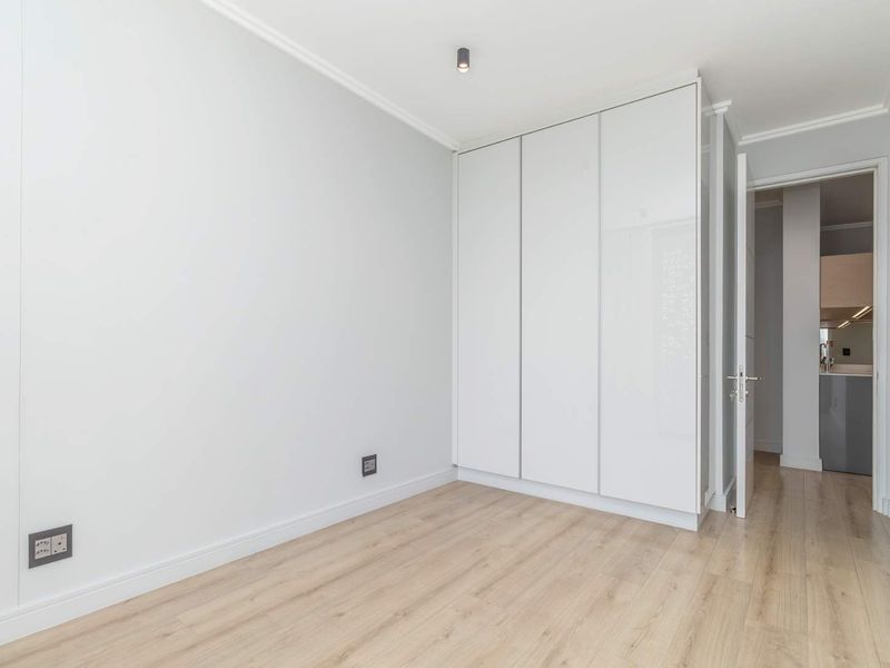A bright, empty bedroom featuring modern laminate flooring, clean white walls, and a large built-in wardrobe with sleek white doors.