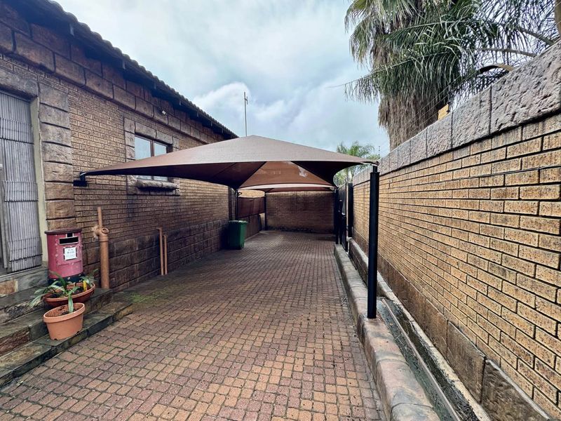 A paved driveway area featuring a shade sail carport structure, enclosed by face brick boundary walls.