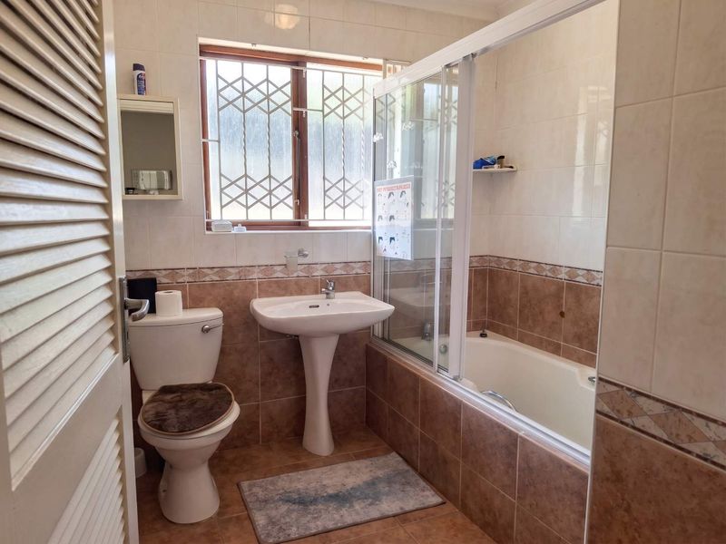 A functional bathroom featuring a pedestal basin, toilet, and a combined bathtub with a shower screen, finished with neutral wall and floor tiling.