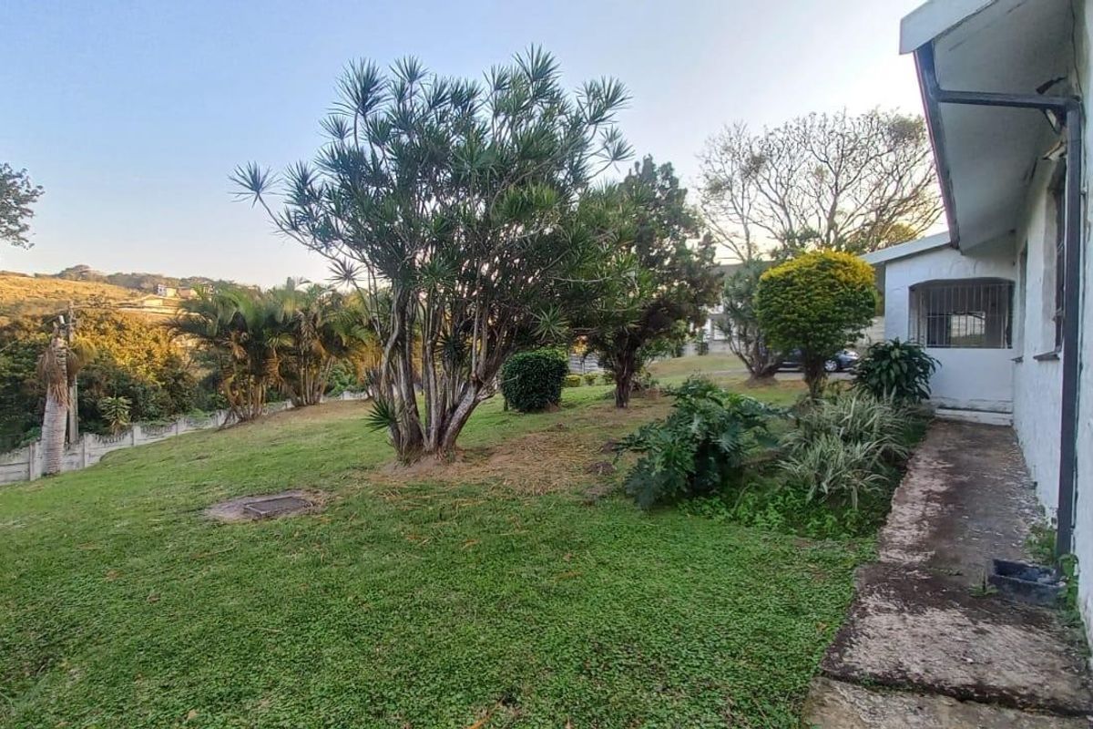A view of the rear garden area featuring a lawn, mature trees, and a concrete pathway adjacent to the house exterior.