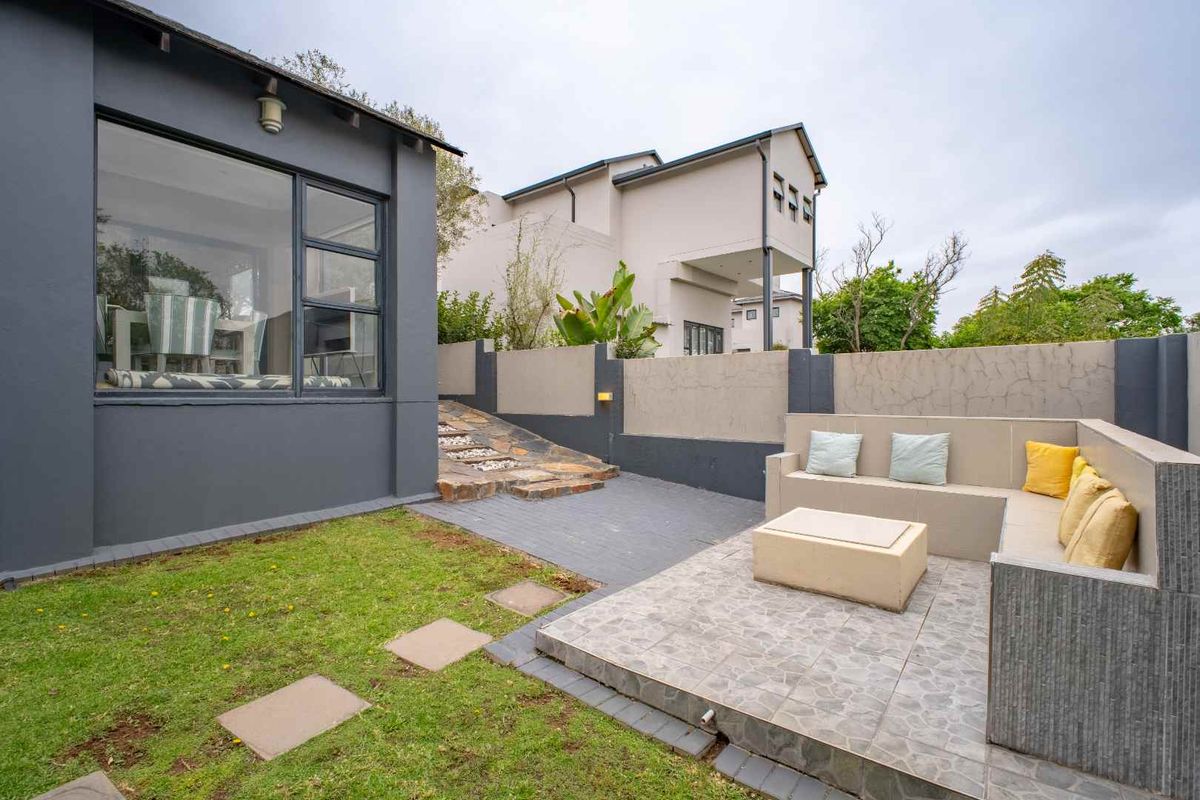 A private rear garden featuring a paved patio area with built-in seating, a small lawn, and stone pathway leading to the main house structure.