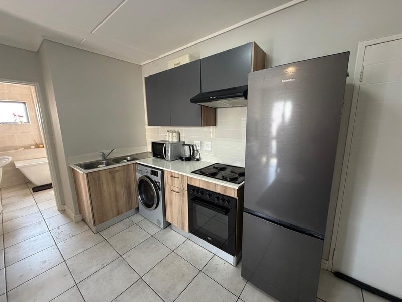 Compact modern kitchen featuring wood-finish cabinetry, integrated appliances, tiled backsplash, and durable floor tiles, with an adjacent view into the bathroom.