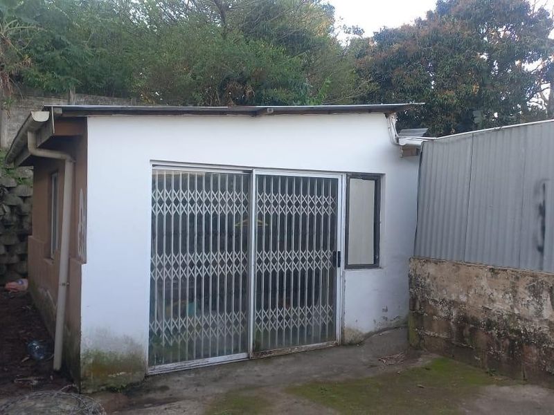 Rear view of a detached outbuilding featuring a sliding security gate, concrete retaining wall, and weathered exterior finishes requiring maintenance.