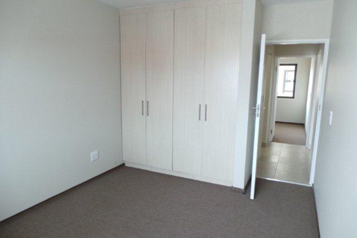 A clean, empty bedroom featuring neutral carpeting and built-in light wood-finish cupboards, with a doorway leading to a tiled hallway.
