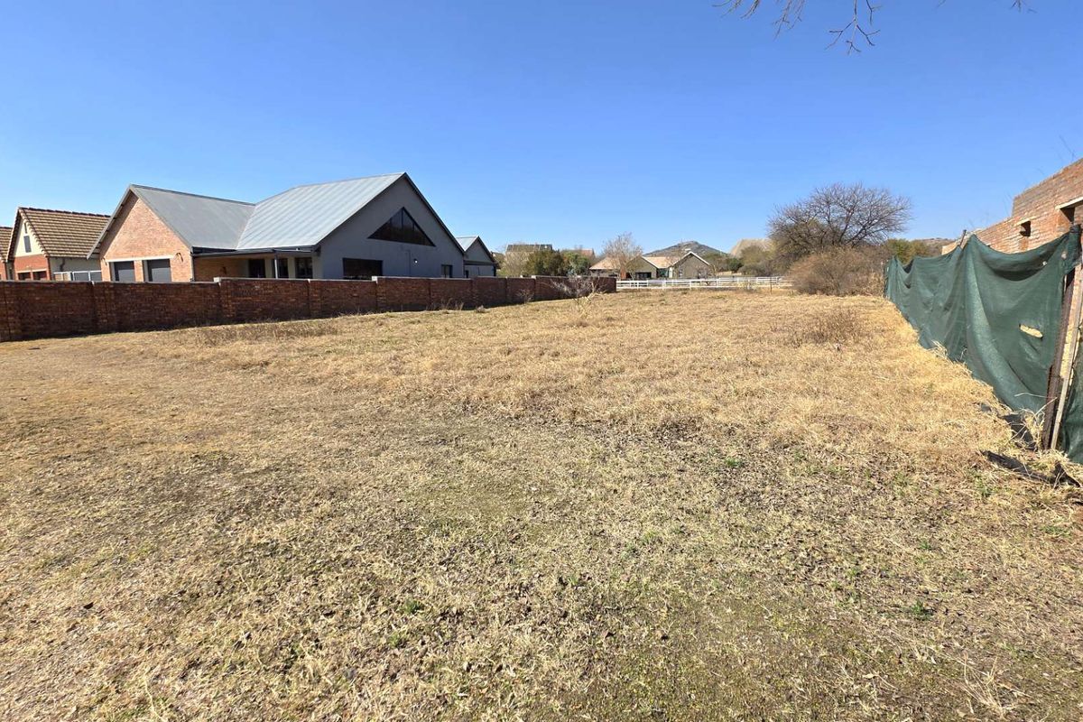 A vacant residential plot featuring dry grass, enclosed by a brick boundary wall and temporary shade cloth fencing.