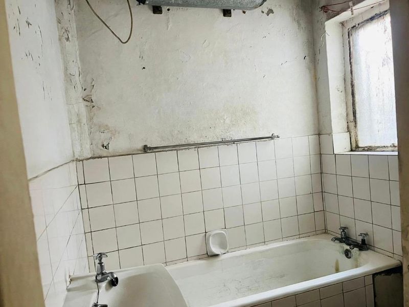 A dated bathroom requiring significant renovation, featuring a standard bathtub, tiled splashback, and visible wall surface deterioration.