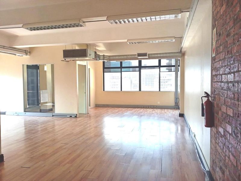 Spacious open-plan office area featuring laminate flooring, exposed brick accents, industrial-style exposed ducting, and large windows providing ample natural light.