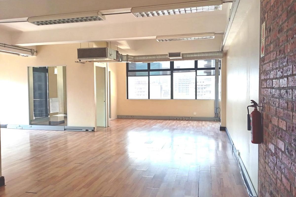 Spacious open-plan office area featuring laminate flooring, exposed brick accents, industrial-style exposed ducting, and large windows providing ample natural light.