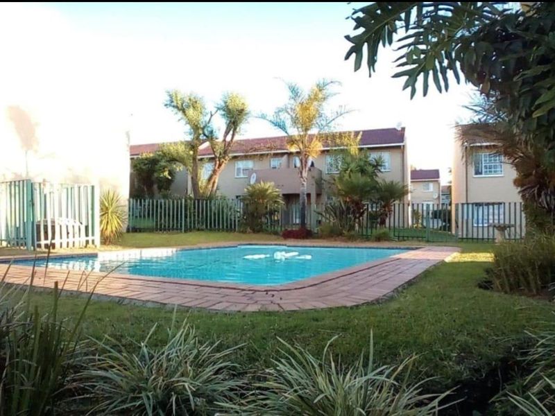 A well-maintained communal swimming pool area featuring brick coping, surrounding lawn, and perimeter fencing within an apartment complex setting.
