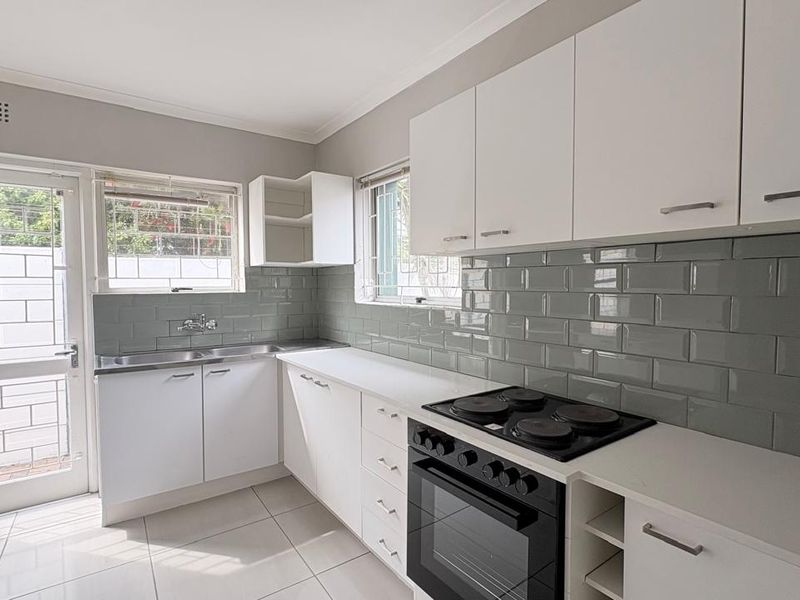 A clean, modern kitchen featuring white cabinetry, a grey subway tile backsplash, built-in oven with electric hob, and direct access to the exterior.