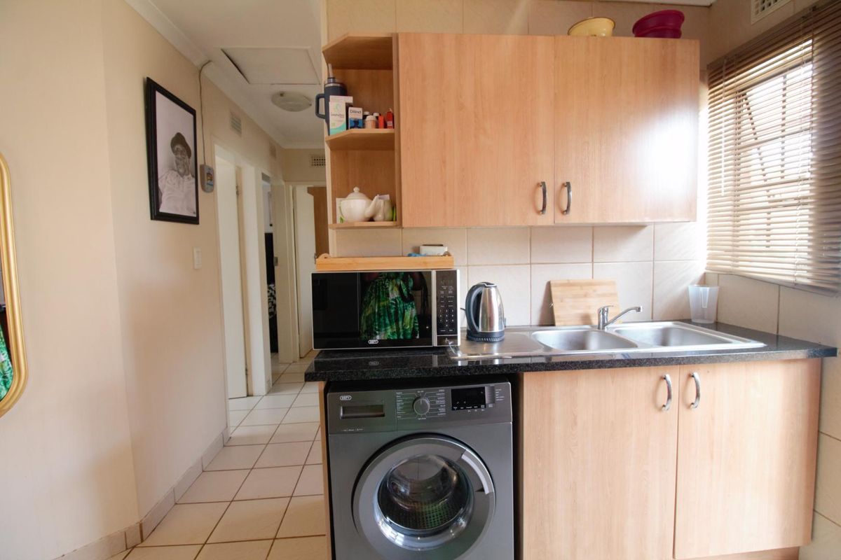 A functional kitchen area featuring light wood cabinetry, granite countertops, a double sink, and tiled flooring, with an integrated space for a washing machine.