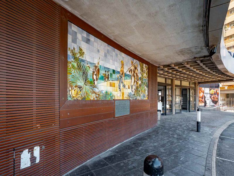Exterior front entrance area featuring a decorative tiled mural, timber slat wall cladding, and a covered concrete walkway with stone paving.