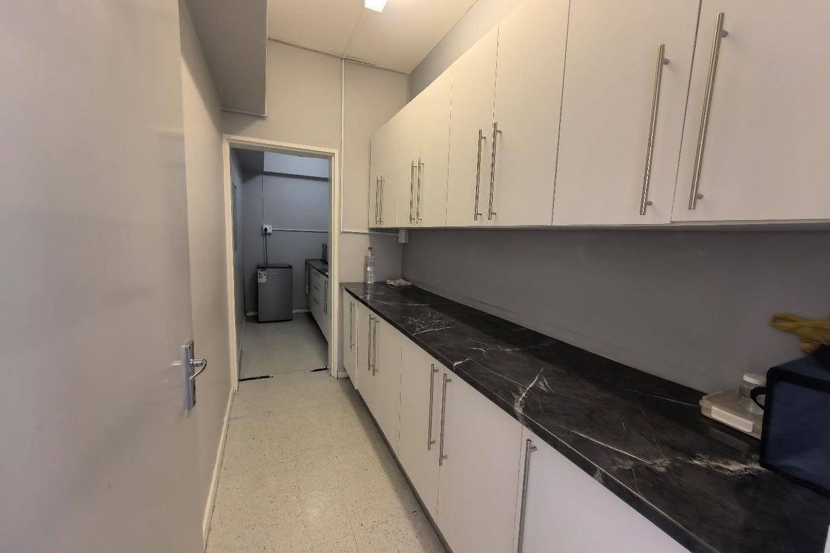 A functional office kitchenette featuring white cabinetry, dark marble-effect countertops, and overhead storage, leading into a secondary utility or prep area.