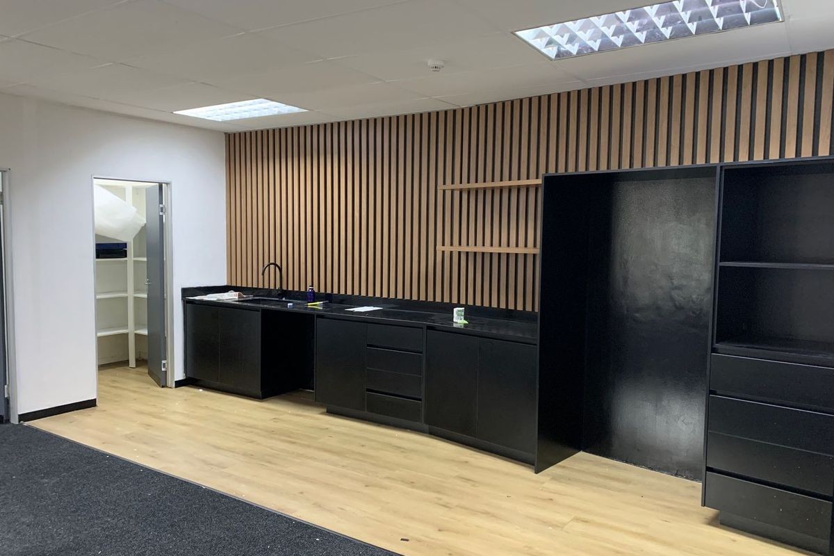 Modern office kitchenette featuring sleek black cabinetry, a contrasting timber slat feature wall, and integrated storage space with a dedicated pantry area.