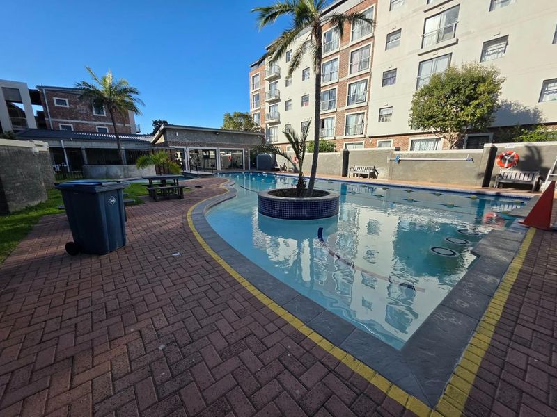 Communal swimming pool area featuring brick paving, a central pool island, and surrounding boundary walls within an apartment complex.