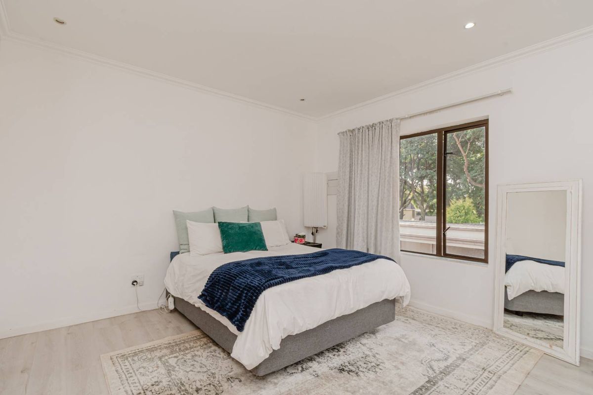 A bright and neutral bedroom featuring laminate flooring, recessed downlighting, and a large window with views of surrounding greenery.