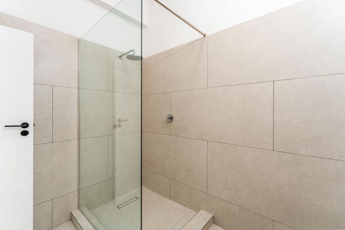 A modern walk-in shower featuring large-format beige wall tiles, a frameless glass screen, and a contemporary rainfall showerhead.