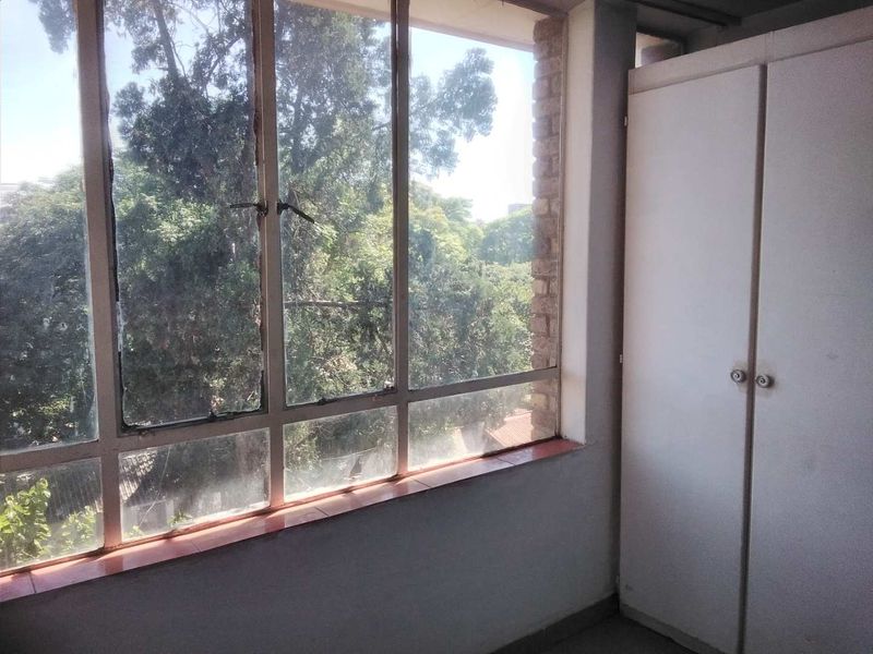 A bright, empty bedroom featuring a built-in wardrobe and large windows with a leafy green outlook.