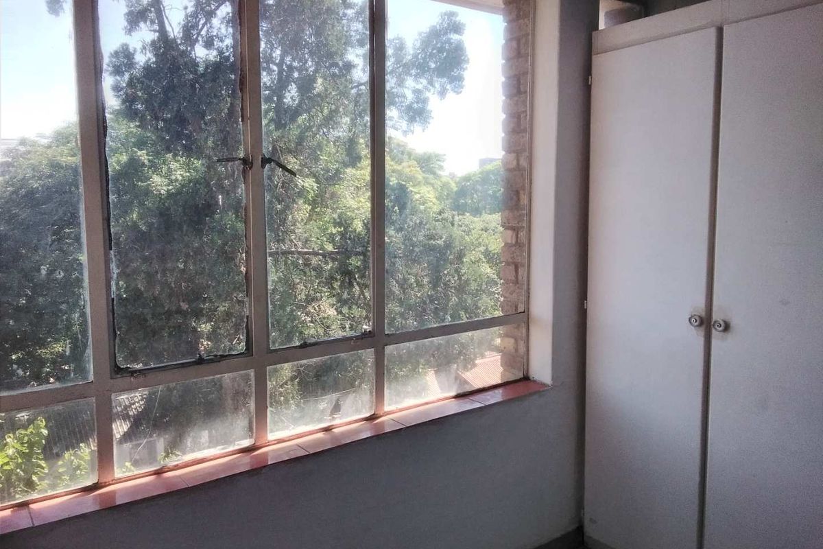 A bright, empty bedroom featuring a built-in wardrobe and large windows with a leafy green outlook.