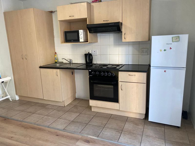 Compact kitchen featuring light wood-tone cabinetry, black countertops, integrated oven and hob, tiled backsplash, and durable ceramic flooring.