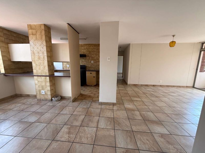An open-plan living and kitchen area featuring tiled flooring, a structural pillar, and dated kitchen cabinetry with a breakfast counter.