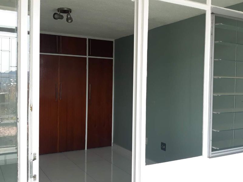 An empty bedroom featuring built-in wooden cupboards, tiled flooring, and louvered windows, viewed from an adjacent balcony area.