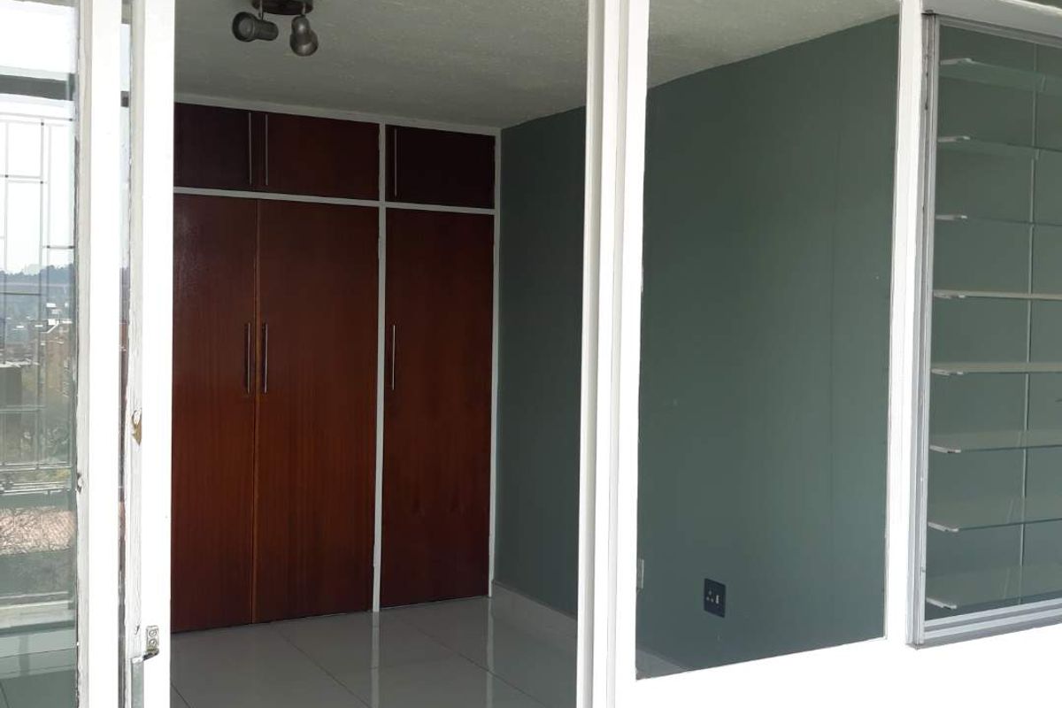 An empty bedroom featuring built-in wooden cupboards, tiled flooring, and louvered windows, viewed from an adjacent balcony area.