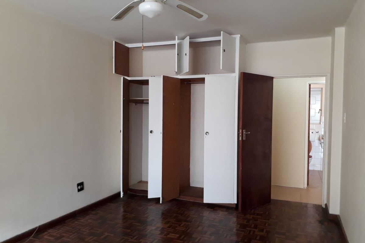 A bedroom featuring classic parquet flooring, a ceiling fan, and built-in storage cupboards, with an adjacent view into a bathroom area.