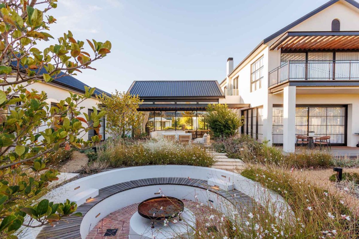 A beautifully landscaped rear garden featuring a sunken circular fire pit area, paved pathways, and a modern multi-level home with a covered patio.