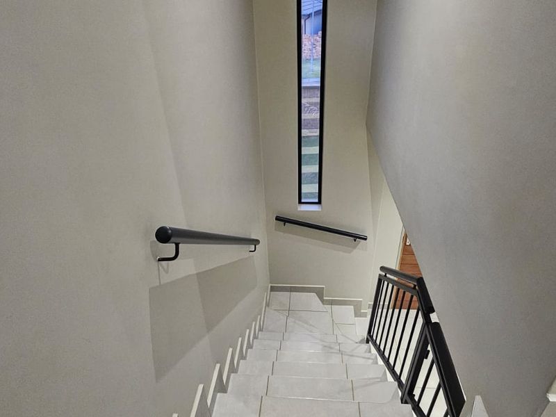 A clean, modern staircase featuring tiled steps, metal handrails, and a tall vertical window providing natural light to the hallway.