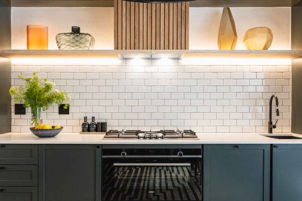 Modern kitchen featuring dark shaker cabinetry, white subway tile backsplash, stone benchtops, integrated gas cooktop, and stylish timber accent rangehood with under-shelf lighting.
