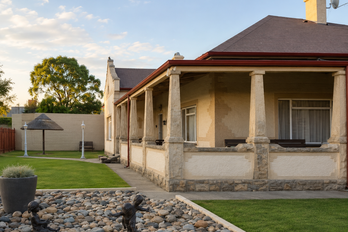 A charming heritage-style home featuring a wrap-around veranda with stone columns, set within a well-maintained garden with a lawn and decorative rockery.